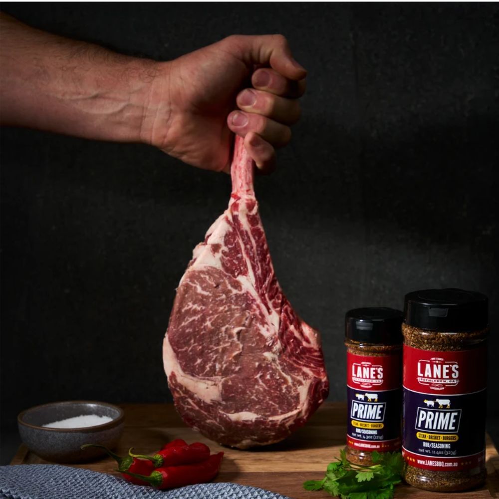 Lane’s Prime BBQ Seasoning small and Pitmaster size beside a raw tomahawk steak, ideal for seasoning beef, brisket, burgers and barbecue meats.