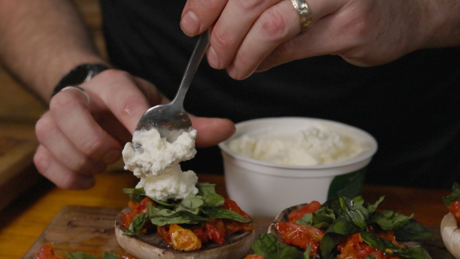 Close-up of a spoon adding ricotta on top of stuffed mushroom caps filled with basil and sun-dried tomatoes.