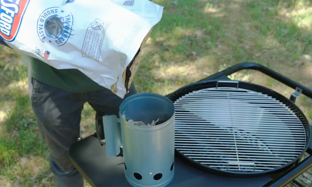 Person pouring charcoal into a chimney starter next to an SNS Master Kettle Grill with the cooking grate in place.