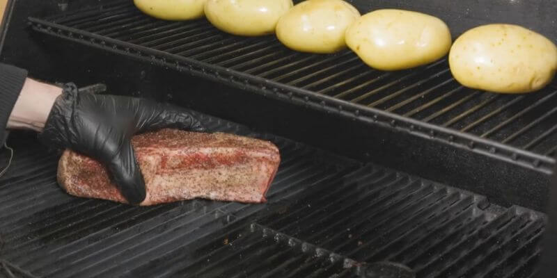 Placing seasoned steak on a Masterbuilt Gravity Feed Smoker grill with whole potatoes cooking on the upper rack.