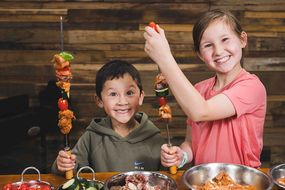 Two smiling children holding skewers with marinated meat and fresh vegetables ready for grilling.