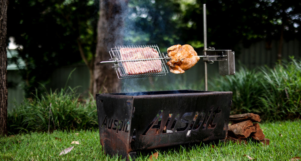 Auspit rotisserie setup outdoors, cooking meat over an open fire pit. A wire basket holds sliced meat alongside a whole seasoned chicken roasting over smouldering wood.