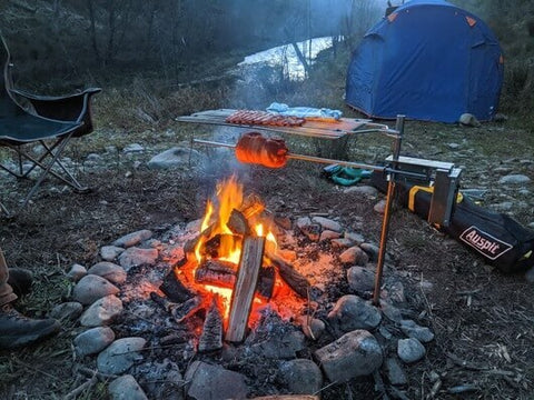 Auspit rotisserie set up over a campfire, cooking meat and sausages on skewers. The fire is surrounded by rocks, with a camping chair and tent in the background near a riverbank.