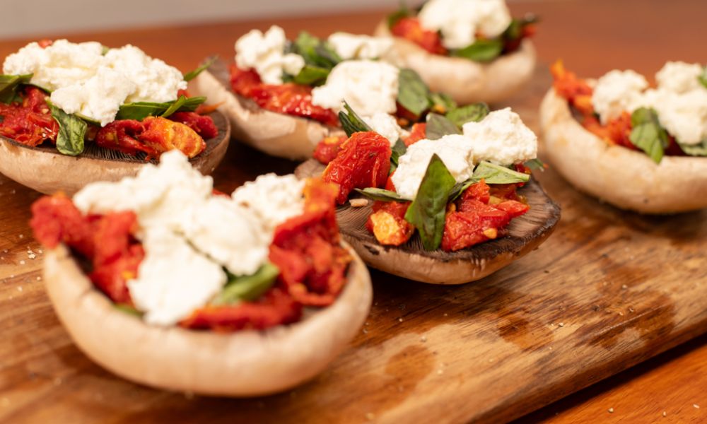 Close-up of stuffed mushroom caps filled with sun-dried tomatoes, fresh basil, and dollops of ricotta on a wooden board before grilling.