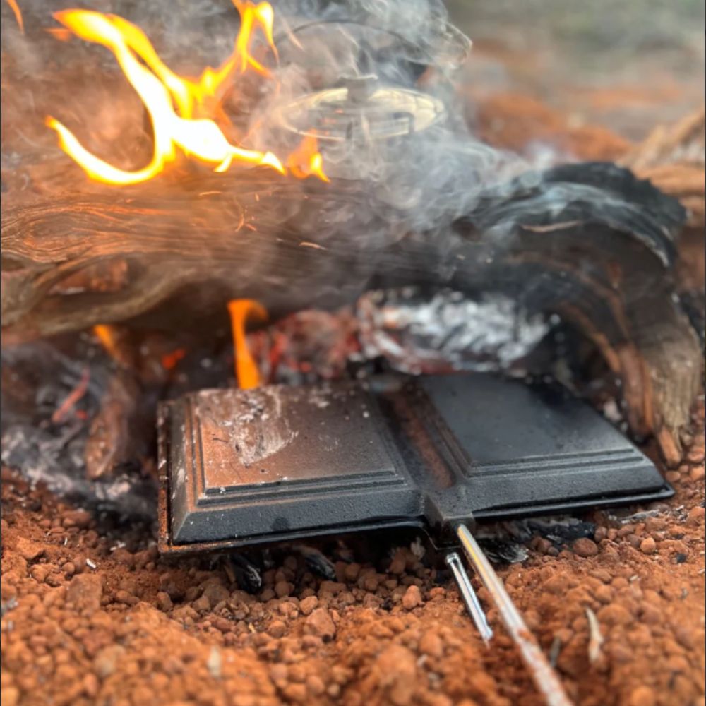 Jumbo Double Jaffle Iron by Wildtrack cooking over an open flame, with smoke rising and heat radiating from the fire.