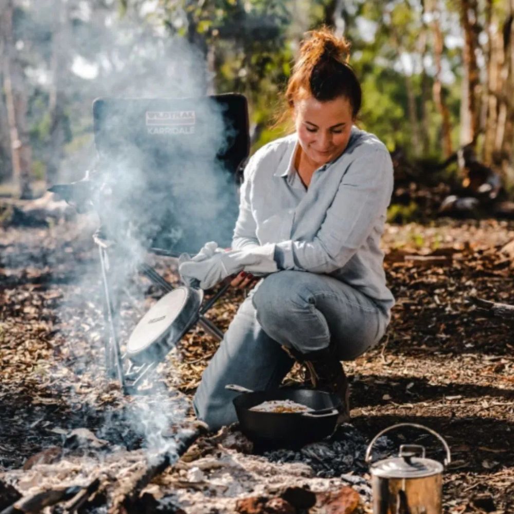 A person cooking over an open fire using a 3.2 Quart Pre-Seasoned Cast Iron 2-in-1 Combo Cooker, with smoke rising from the fire in the background."