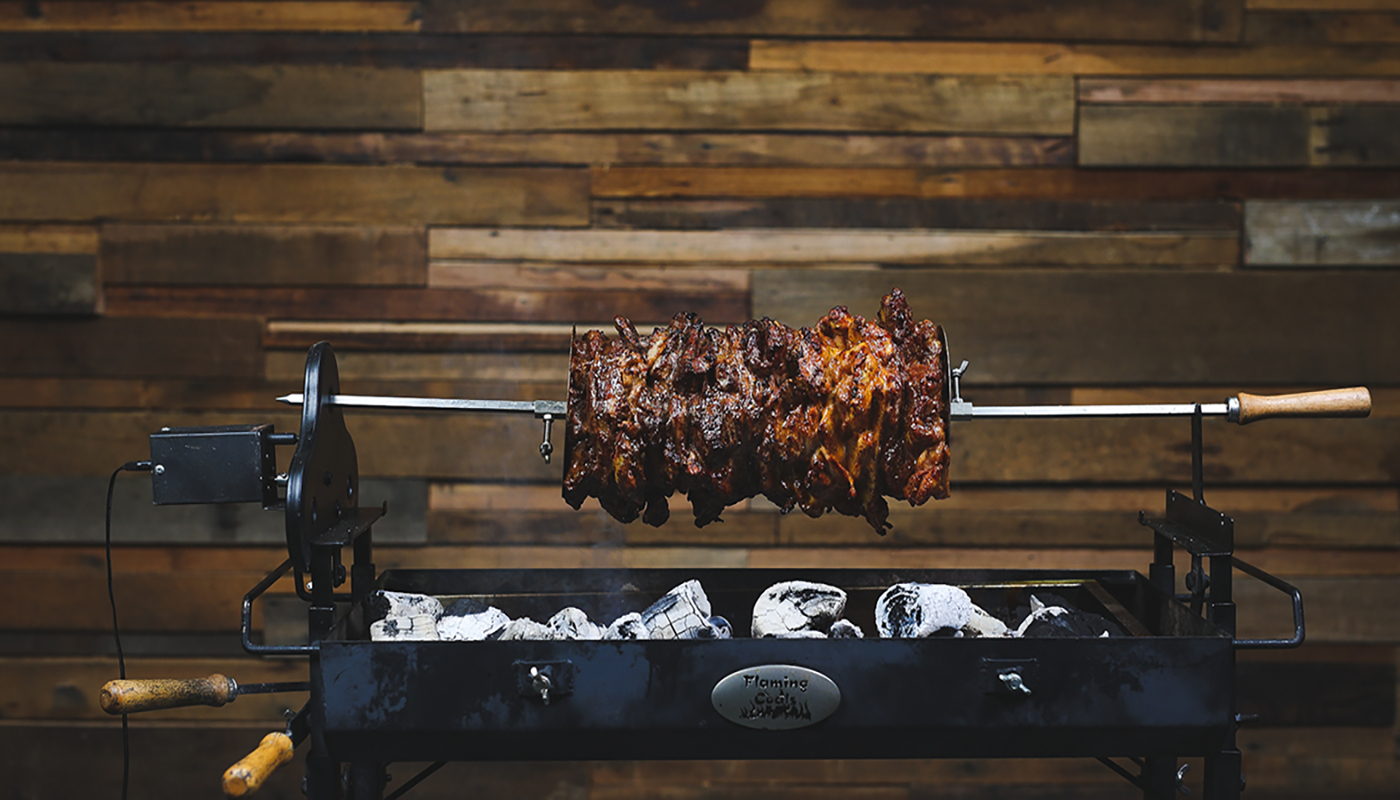 A large skewer of chicken and lamb gyros roasting over hot charcoal on a Flaming Coals rotisserie. The meat is golden and crisp, slowly turning above glowing coals against a wooden backdrop.