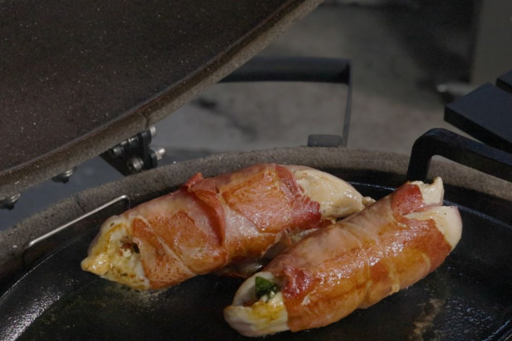 Two prosciutto-wrapped stuffed chicken breasts sizzling in a cast iron pan inside an SNS Kamado smoker, mid-cook with a golden sear developing.