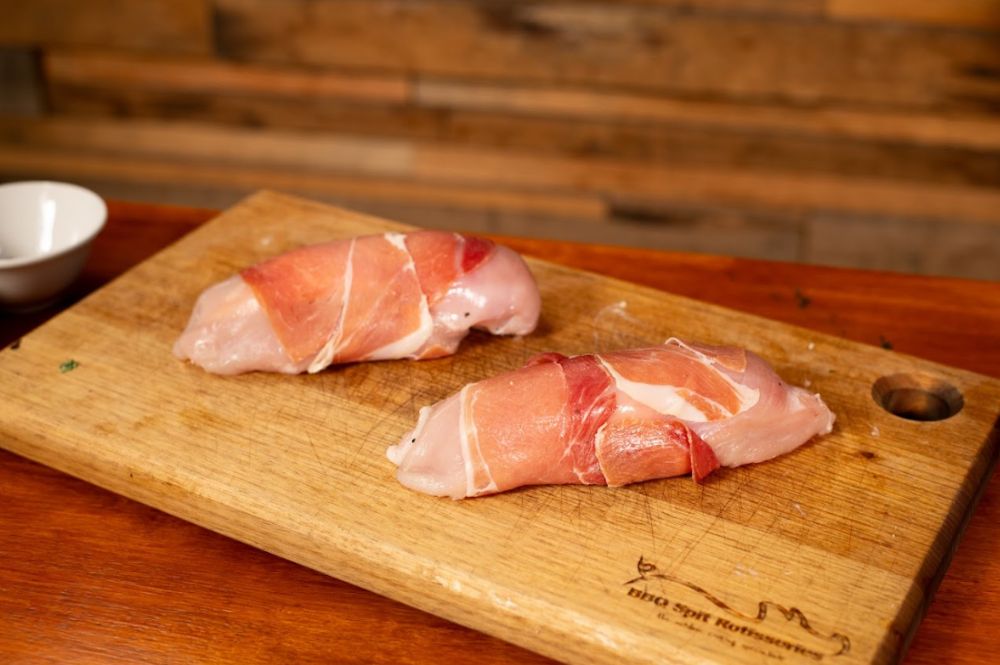 Two raw chicken breasts wrapped in prosciutto, placed on a wooden cutting board, ready to be cooked in the smoker.