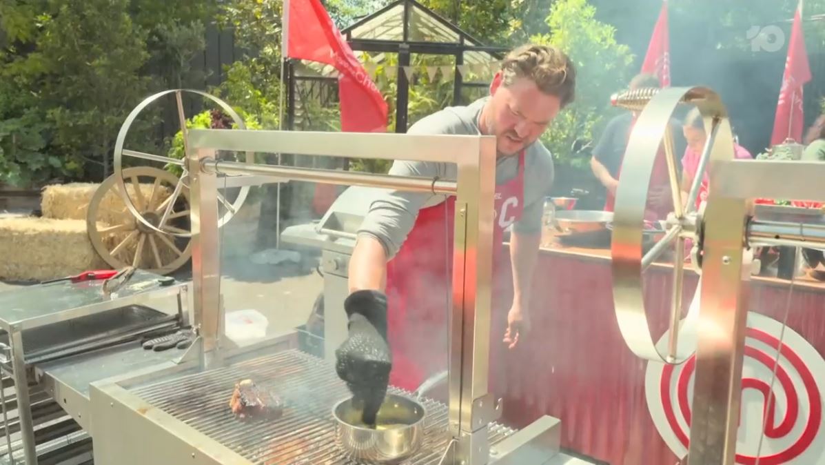 Contestant grilling a steak on a Parrilla grill, applying sauce with a brush  surrounded by smoke, in a MasterChef competition setting