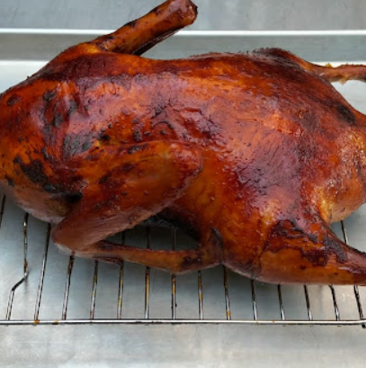 Whole roasted duck with dark golden, crispy skin resting on a wire rack over a metal tray after being cooked in a wood fired pizza oven.”