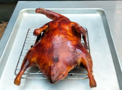 Whole duck cooked in a wood fired pizza oven, resting on a wire rack over a metal tray with crispy golden brown skin.