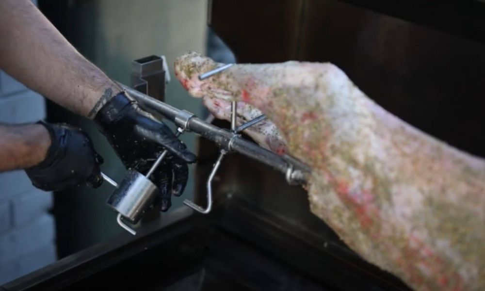 Person wearing black gloves adjusting a counter balance weight on a spit roaster skewer holding a whole lamb.