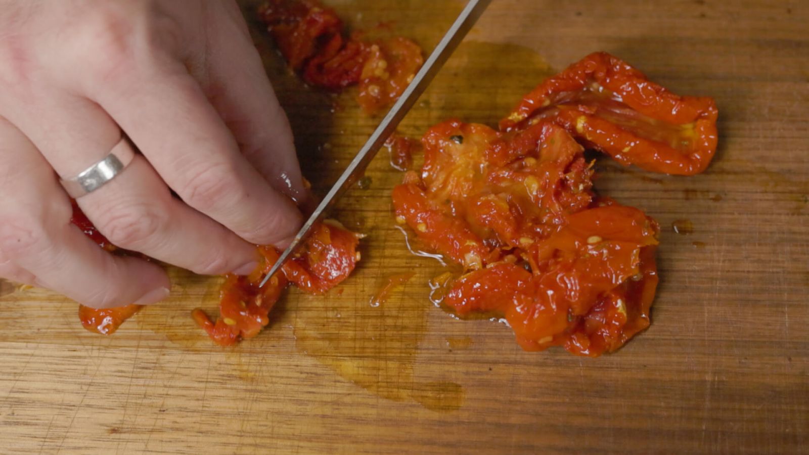 Close-up of hands finely chopping sun-dried tomatoes on a wooden board with a sharp knife.
