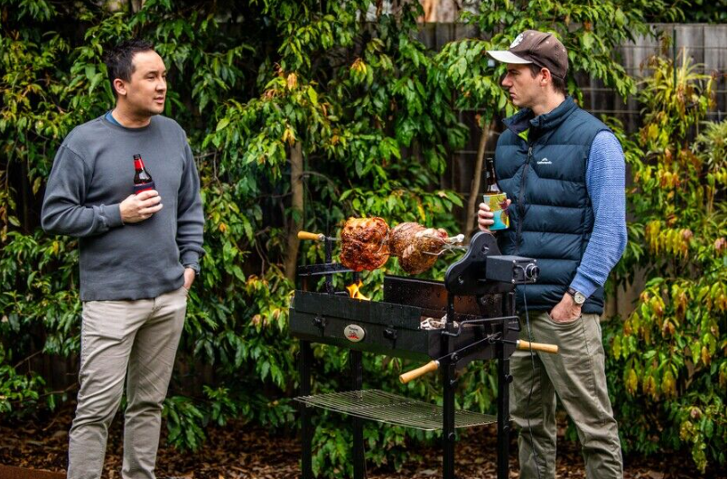 Two men standing beside a Flaming Coals Cyprus spit roaster cooking rotisserie meats in a backyard.