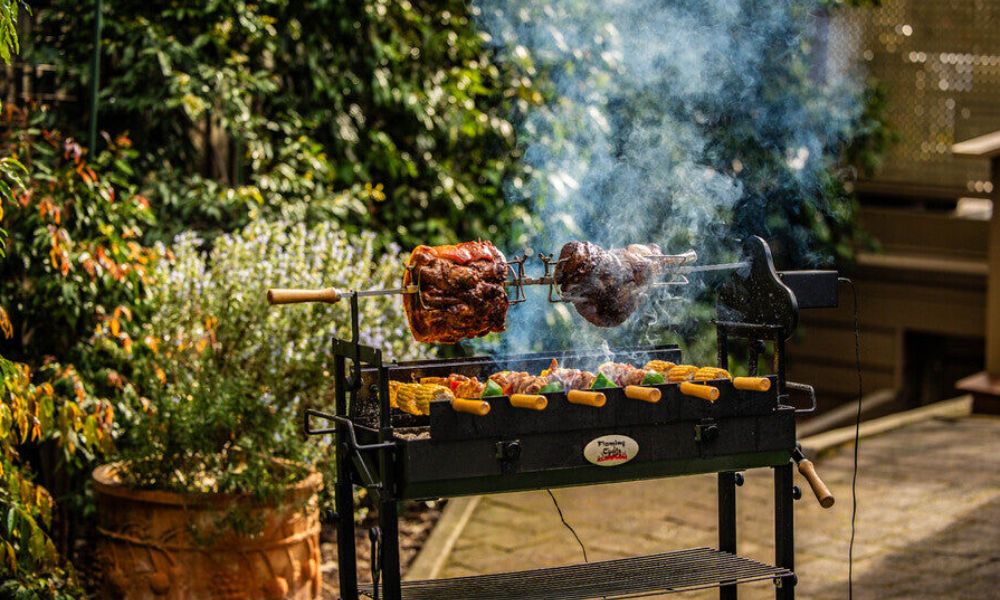 Outdoor charcoal spit roaster cooking two large pieces of meat on the rotisserie with corn and vegetables grilling below, surrounded by smoke and garden greenery.