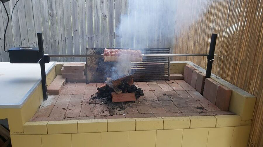DIY spit rotisserie setup with a motorized spit over a bed of hot coals, cooking a rack of meat. The structure is built with bricks and surrounded by a wooden fence.
