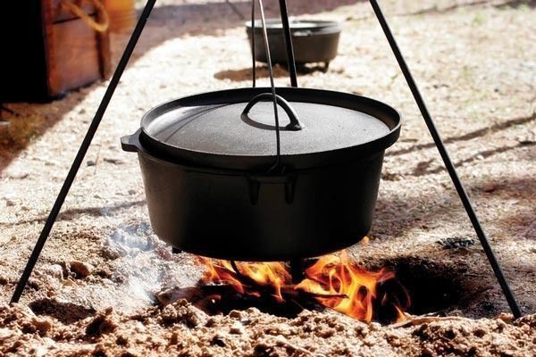 A 12 Quart Dutch Oven (11.3L) hanging over a campfire, suspended by a tripod, with flames visible below, showcasing rustic outdoor cooking.