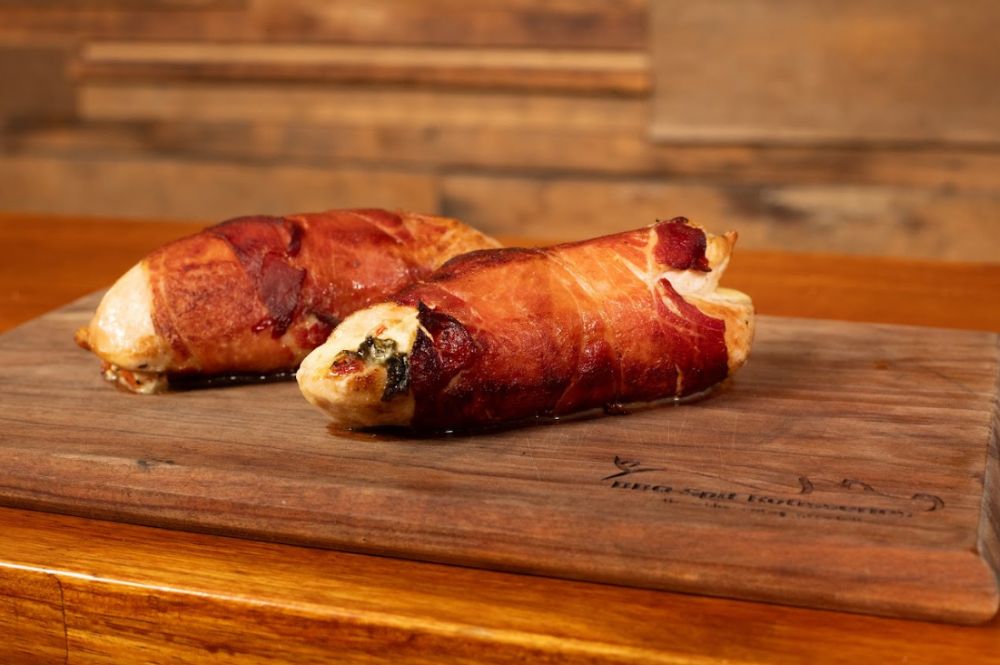 Chicken breast wrapped in crispy prosciutto, served on a rustic wooden board, showcasing the golden-brown, juicy exterior with hints of melted cheese and herbs peeking out.