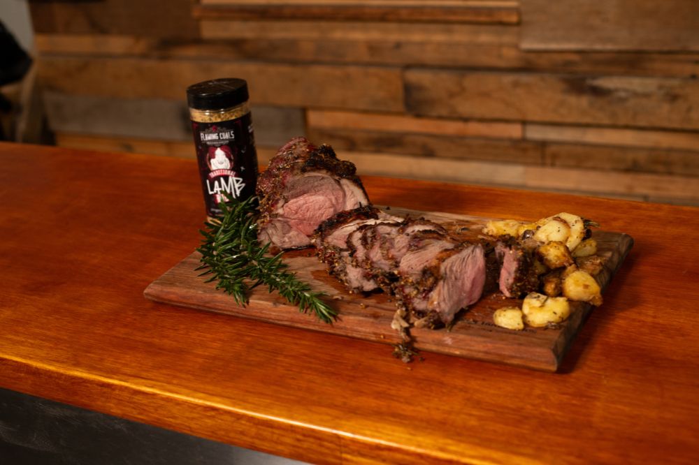  Roast lamb seasoned with Flaming Coals Lamb Rub, sliced and served on a wooden board with golden roasted potatoes and fresh rosemary.