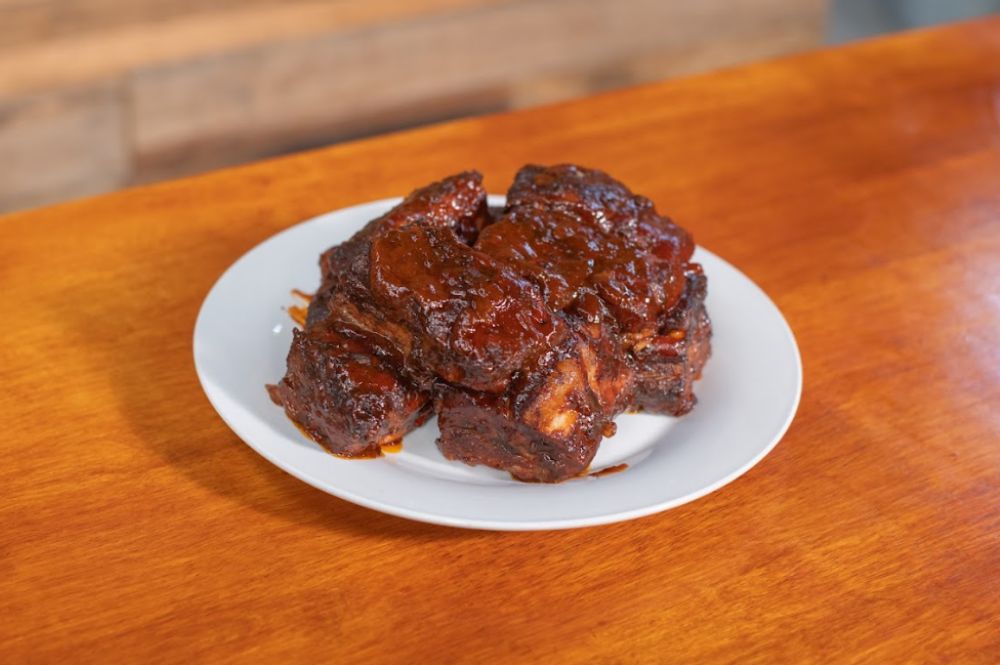 Smoked pork belly bites stacked on a white dish, served on a wooden table. The pork is dark, caramelized, and glistening with a sticky barbecue glaze.
