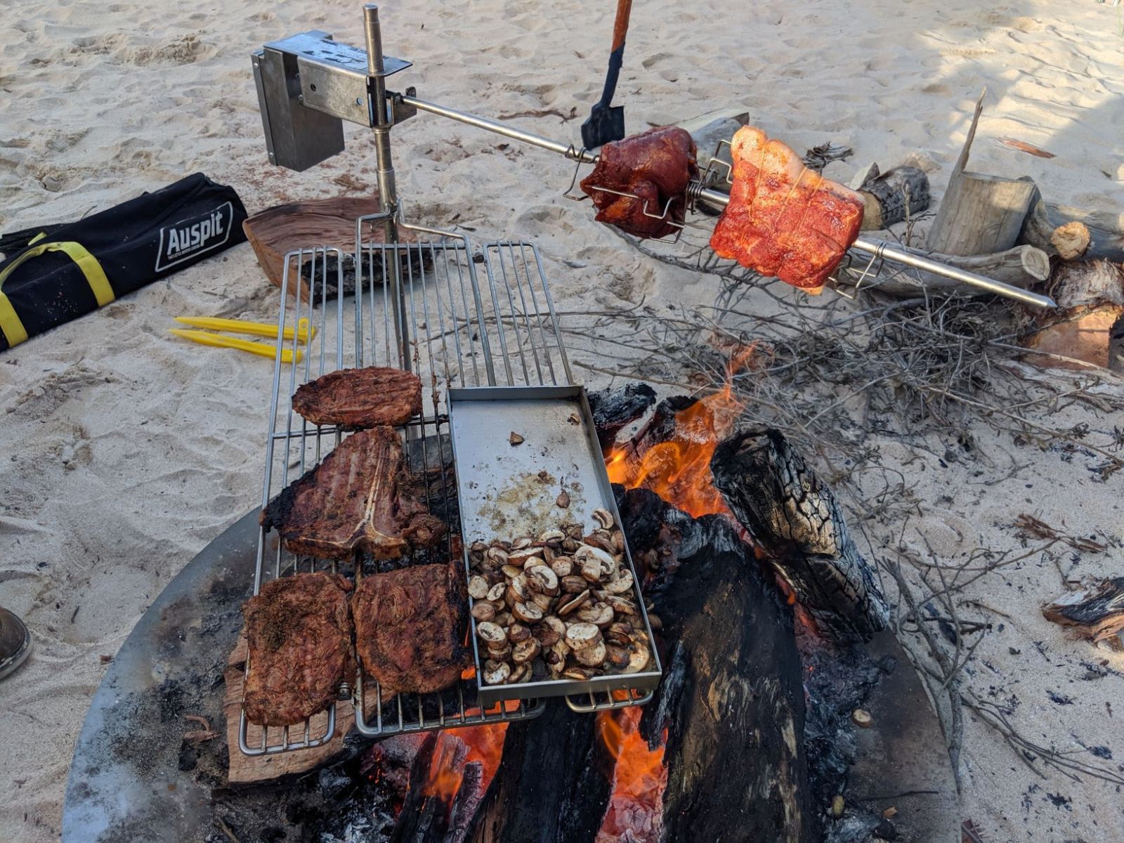 This image show the Deluxe Campfire Swinging Grill cooking steaks on the grill, mushrooms on the hotplate and a spit roast cooking over a fire while camping
