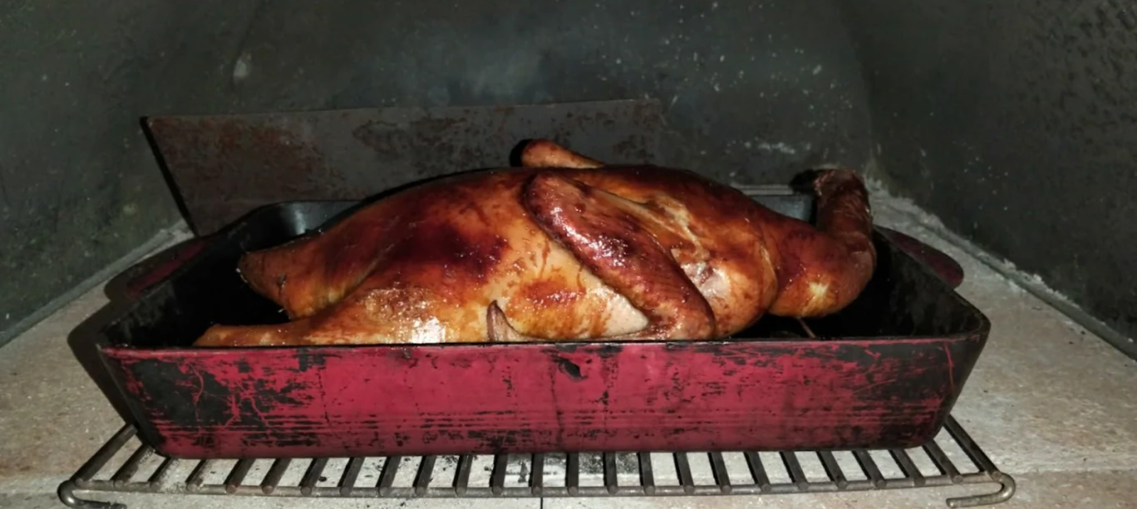 Whole roasted duck with crispy golden-brown skin cooking inside a wood fired pizza oven, placed in a red roasting pan on a metal rack.
