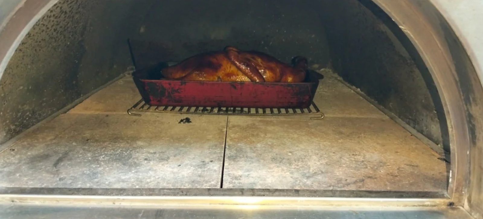 Whole roasted duck with crispy golden skin inside a red roasting pan, placed on a metal rack while cooking in a wood fired pizza oven.
