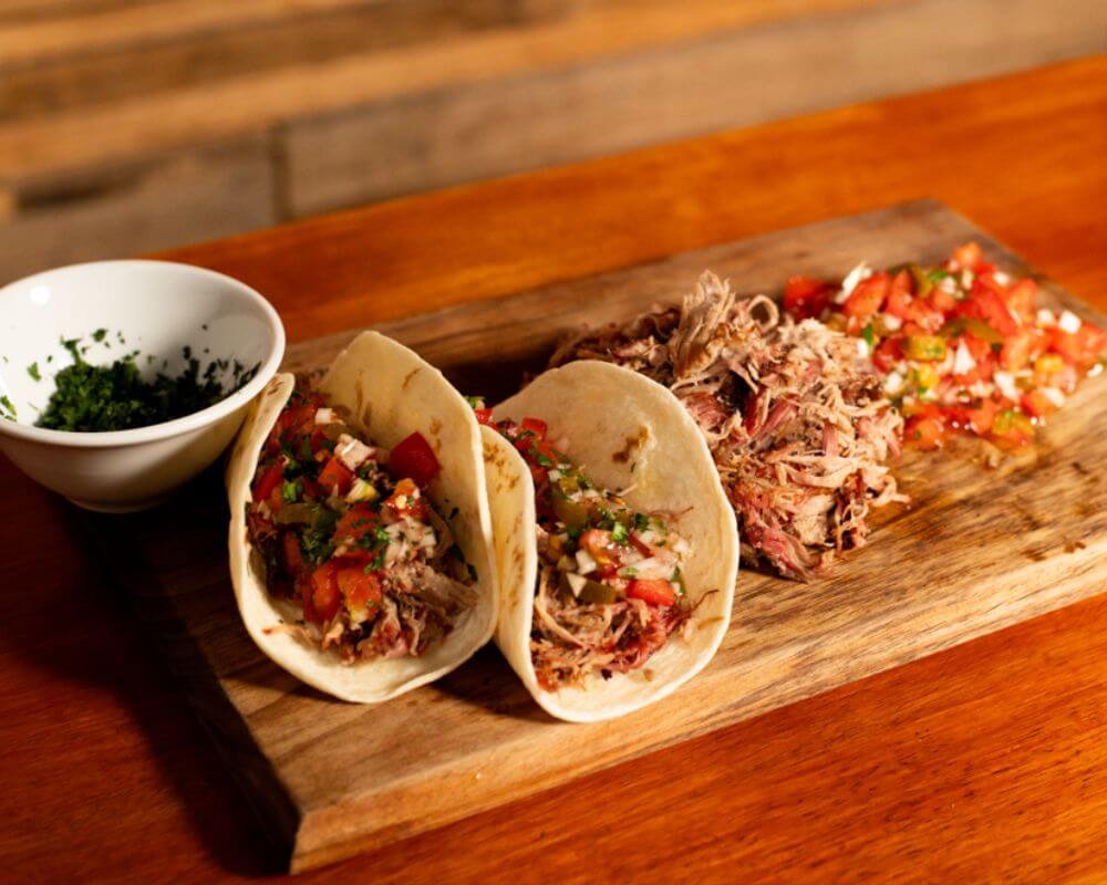 Soft tacos filled with leftover BBQ meat, fresh salsa, and herbs, served on a wooden board with extra toppings on the side.