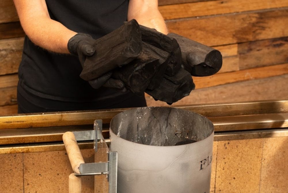 A person wearing black gloves places large pieces of lump charcoal into a charcoal chimney starter positioned on a Flaming Coals Parrilla BBQ.