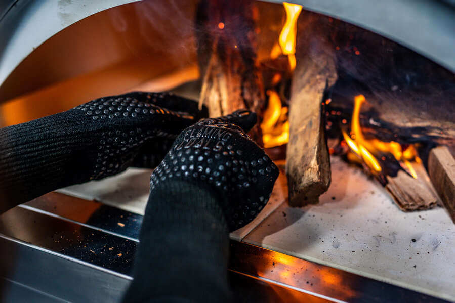 Flaming Coals heat proof gloves adjusting burning firewood inside a pizza oven for safe high-temperature cooking.