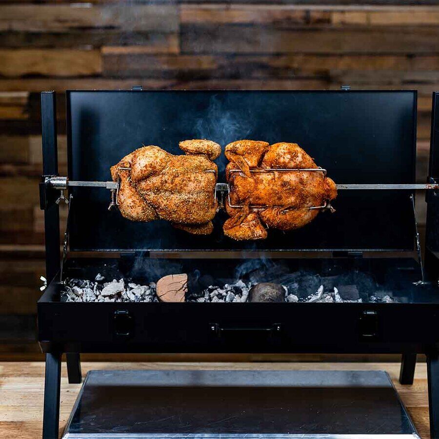 Two seasoned whole chickens cooking on a mini spit rotisserie over hot charcoal.