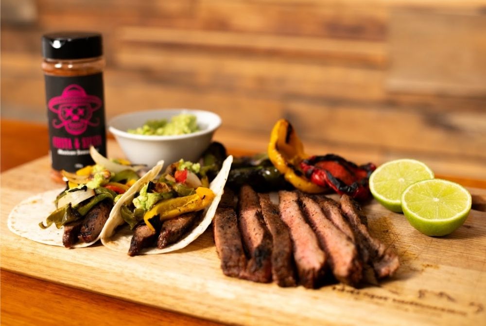  Grilled steak fajitas with tortillas, capsicums, guacamole, lime halves, and BBQ seasoning on a wooden board.