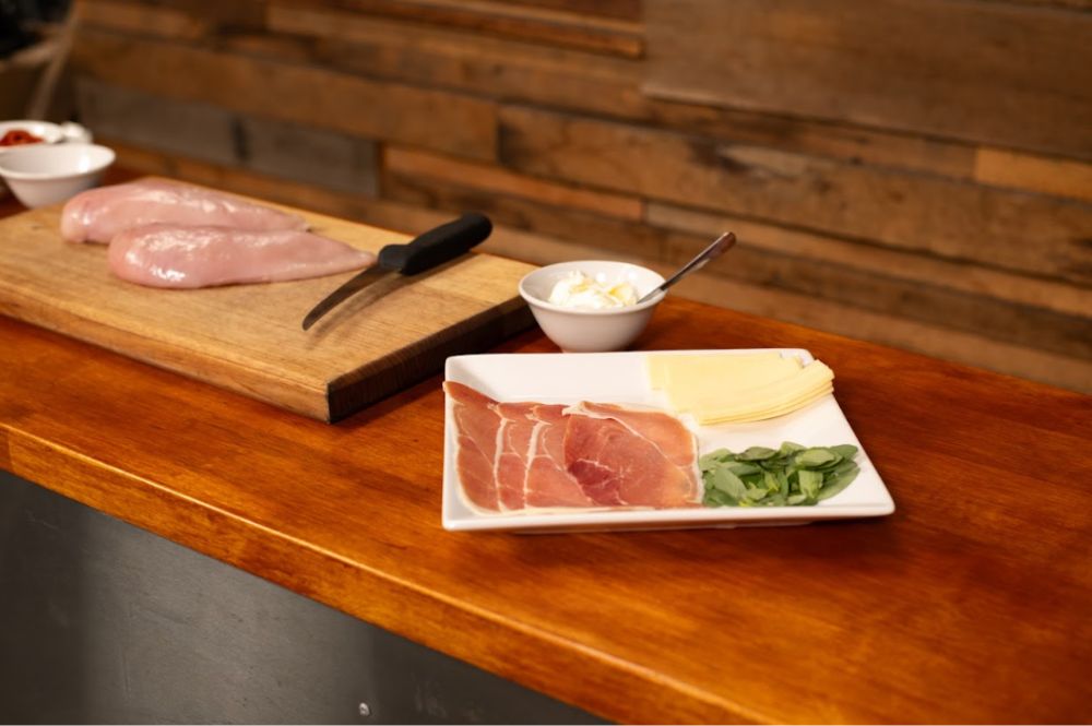 Ingredients for stuffed chicken breast laid out on a wooden countertop, including raw chicken breasts on a cutting board with a knife, and a plate with prosciutto, cheese slices, fresh basil, and a bowl of cream cheese in the background.