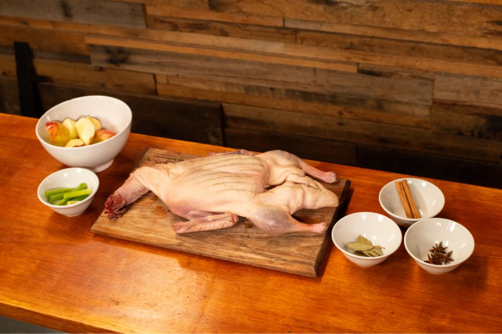 Whole raw duck prepared on a wooden board with bowls of ingredients for spit roasting, including apples, spring onions, bay leaves, cinnamon sticks, and star anise.