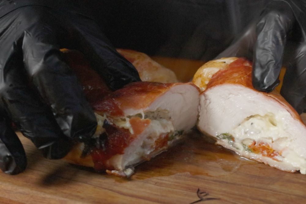 Gloved hands slicing into a juicy, cooked prosciutto-wrapped chicken breast, revealing a melted, cheesy filling with basil and sun-dried tomatoes. Steam rises, showing it's hot and freshly cooked.