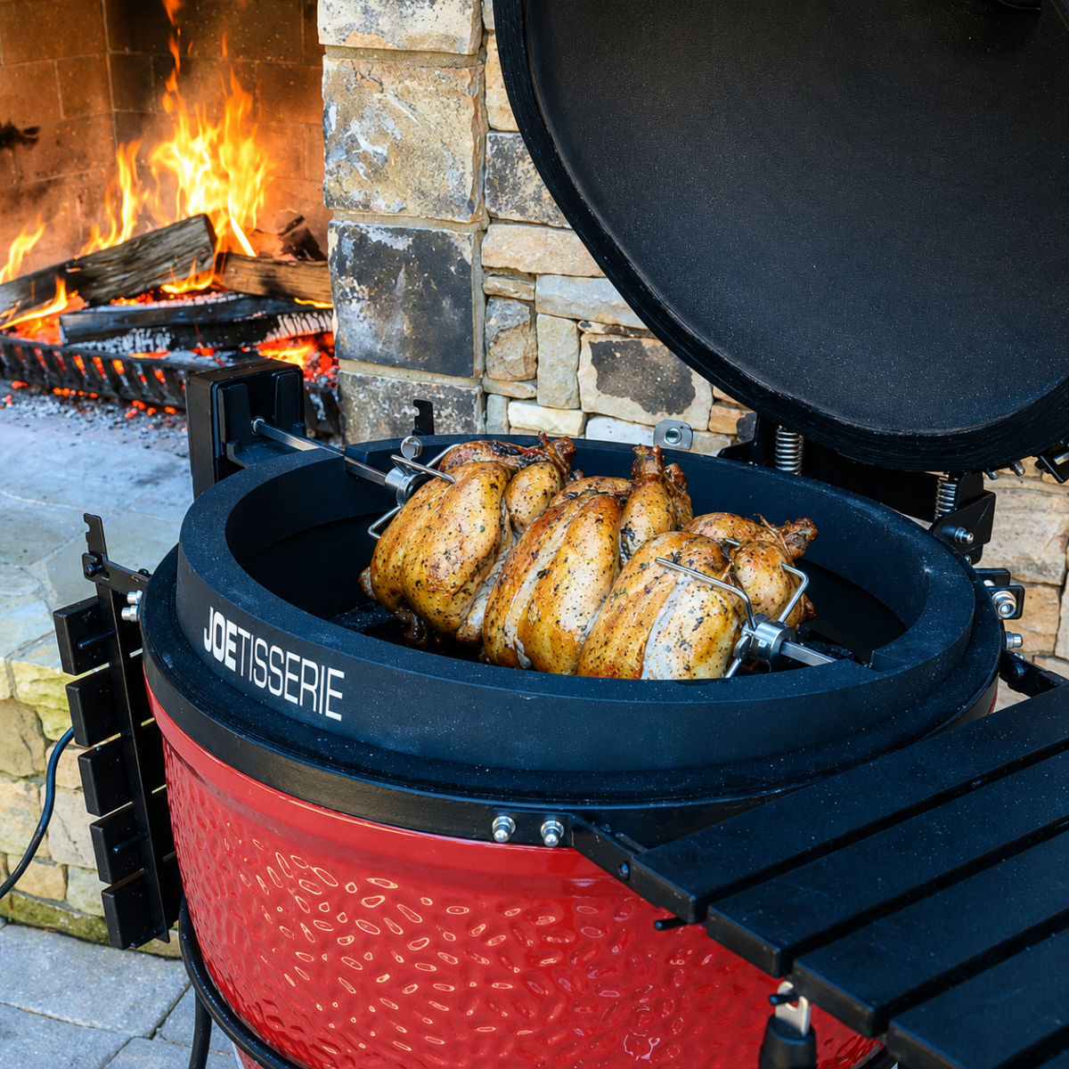 Kamado Joe Big Joe Rotisserie grilling multiple whole chickens on a rotisserie spit with a red ceramic grill in an outdoor backyard setting, showing golden brown cooked chicken and a fire pit in the background.