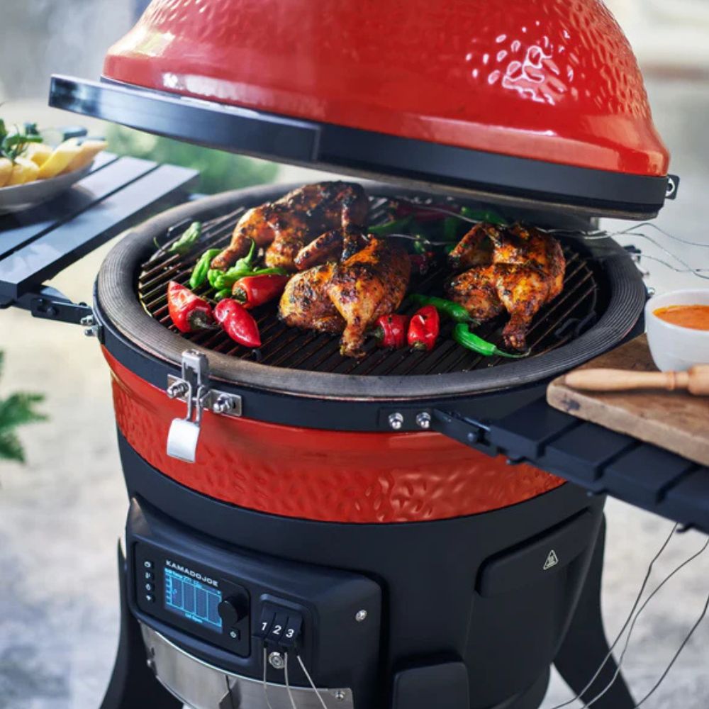 Classic Konnected Digital Charcoal Grill and Smoker by Kamado Joe with red ceramic kamado body, digital control panel, and open lid cooking seasoned chicken and vegetables over charcoal.