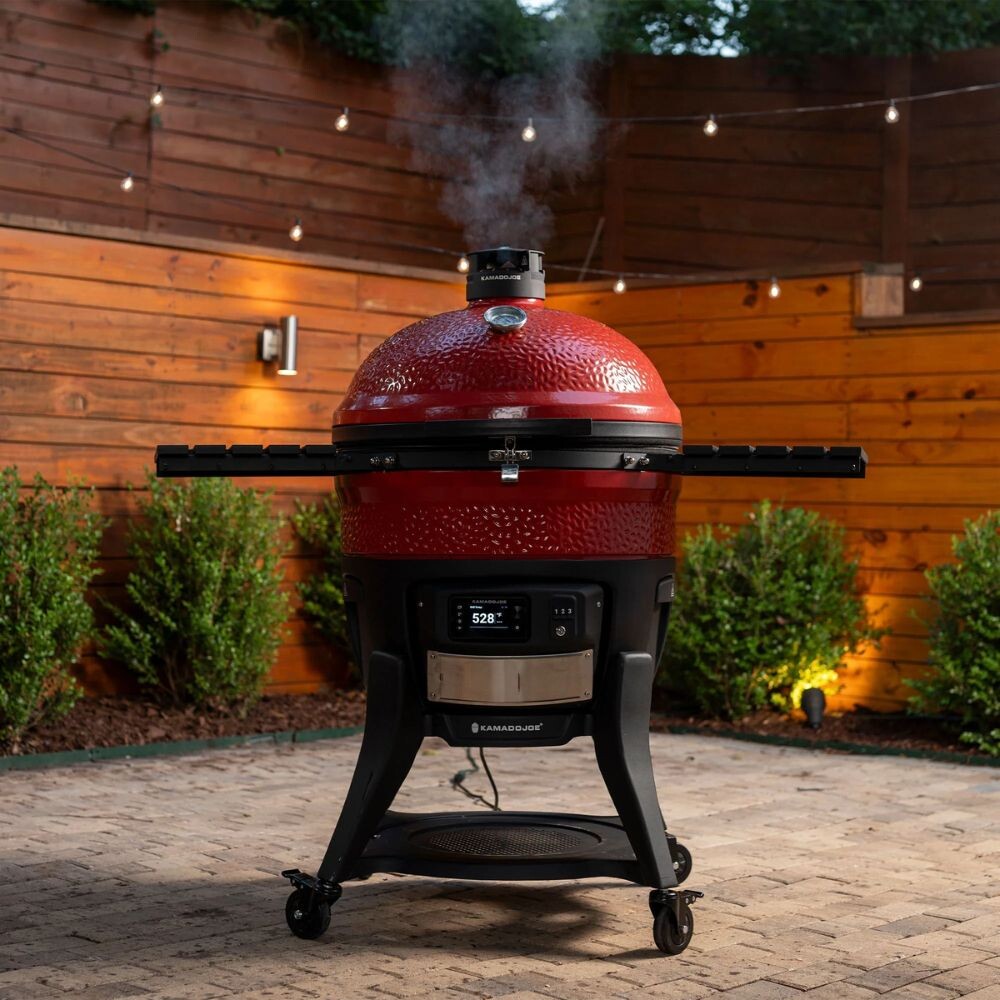 Kamado Joe BBQ Smoker in a backyard setting, showing a red ceramic kamado grill with open vent releasing smoke, digital control panel, and side shelves, ready for charcoal grilling and smoking.
