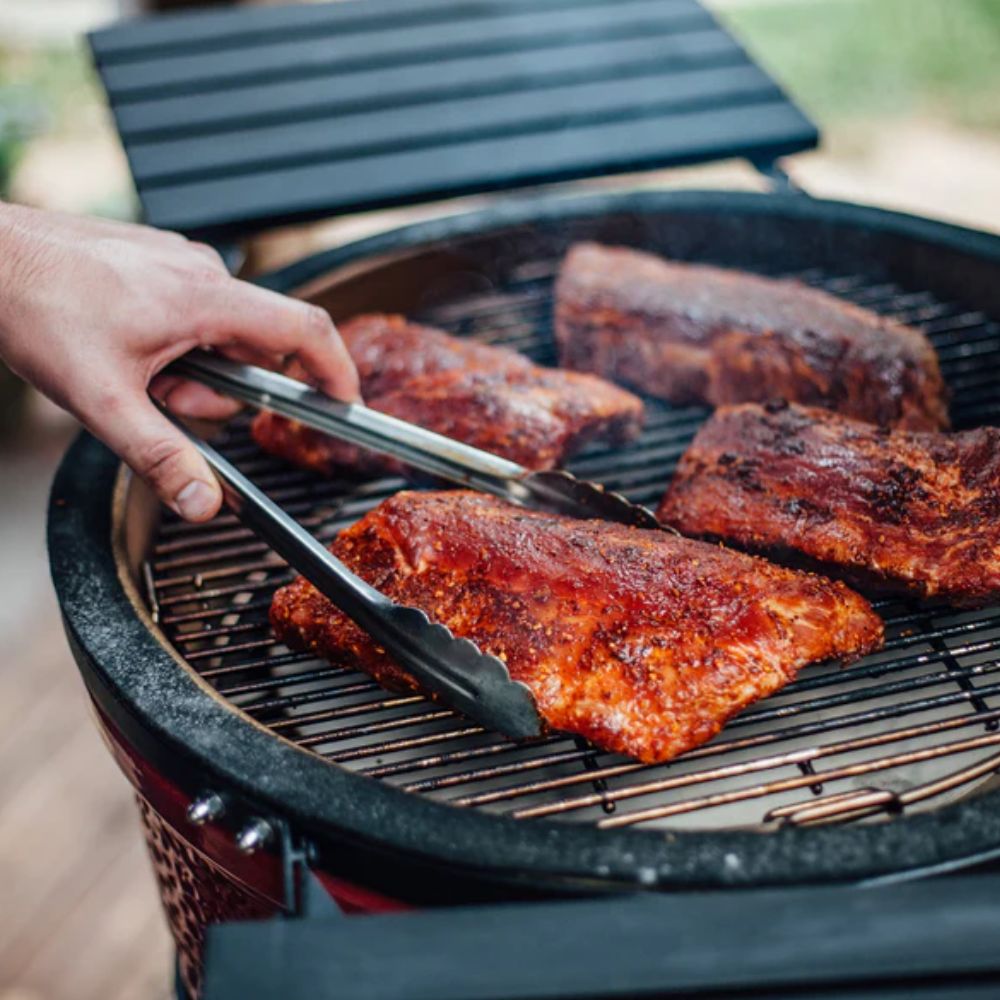 Classic Joe Grill Series I by Kamado Joe cooking seasoned pork ribs on a ceramic charcoal grill with stainless steel tongs and side shelves.