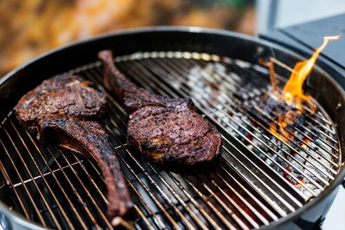 Two tomahawk steaks cooking on a Slow ’N Sear Kettle BBQ using two-zone heat setup with charcoal fire on one side for perfect searing.