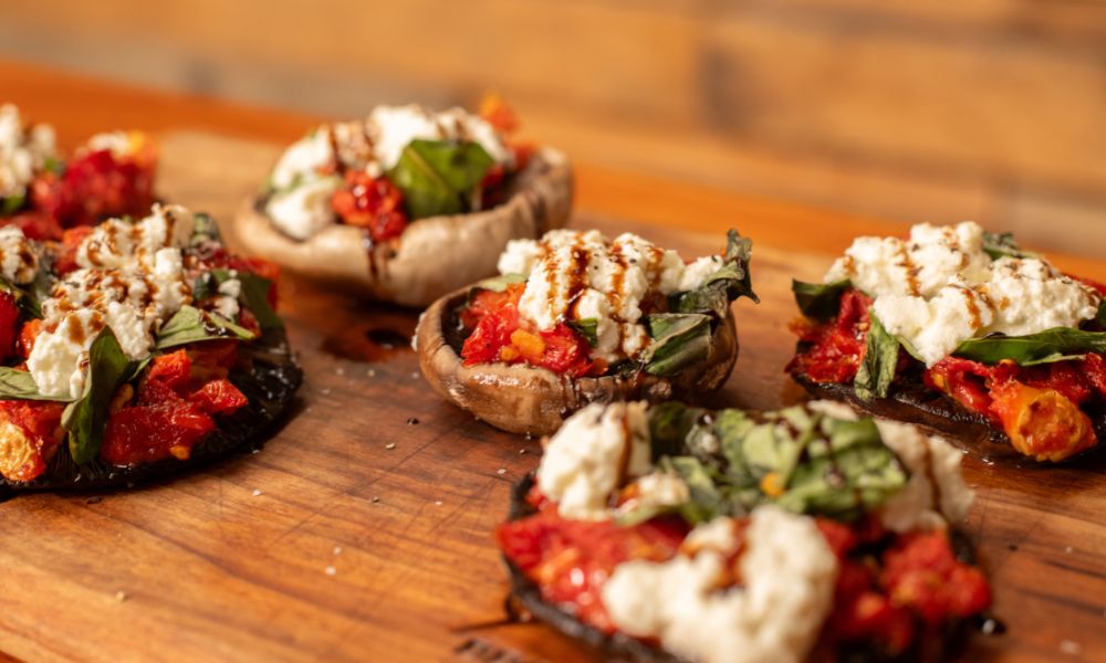 Cooked stuffed mushrooms topped with ricotta, fresh basil, sun-dried tomatoes, and a drizzle of balsamic glaze on a wooden board.