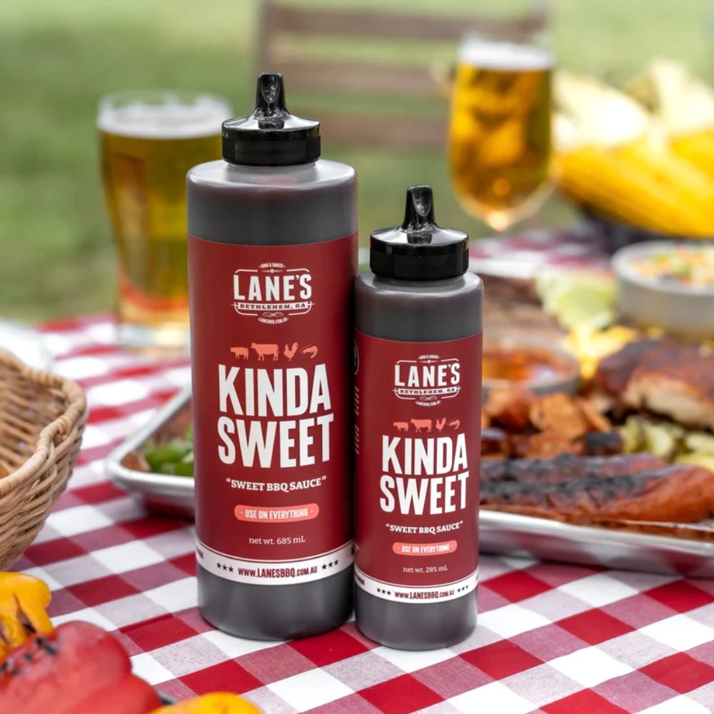 Lane’s BBQ Sauce “Kinda Sweet” displayed on a red checkered table with grilled chicken wings, corn, and coleslaw, showcasing a sweet and tangy barbecue sauce served with classic BBQ sides.