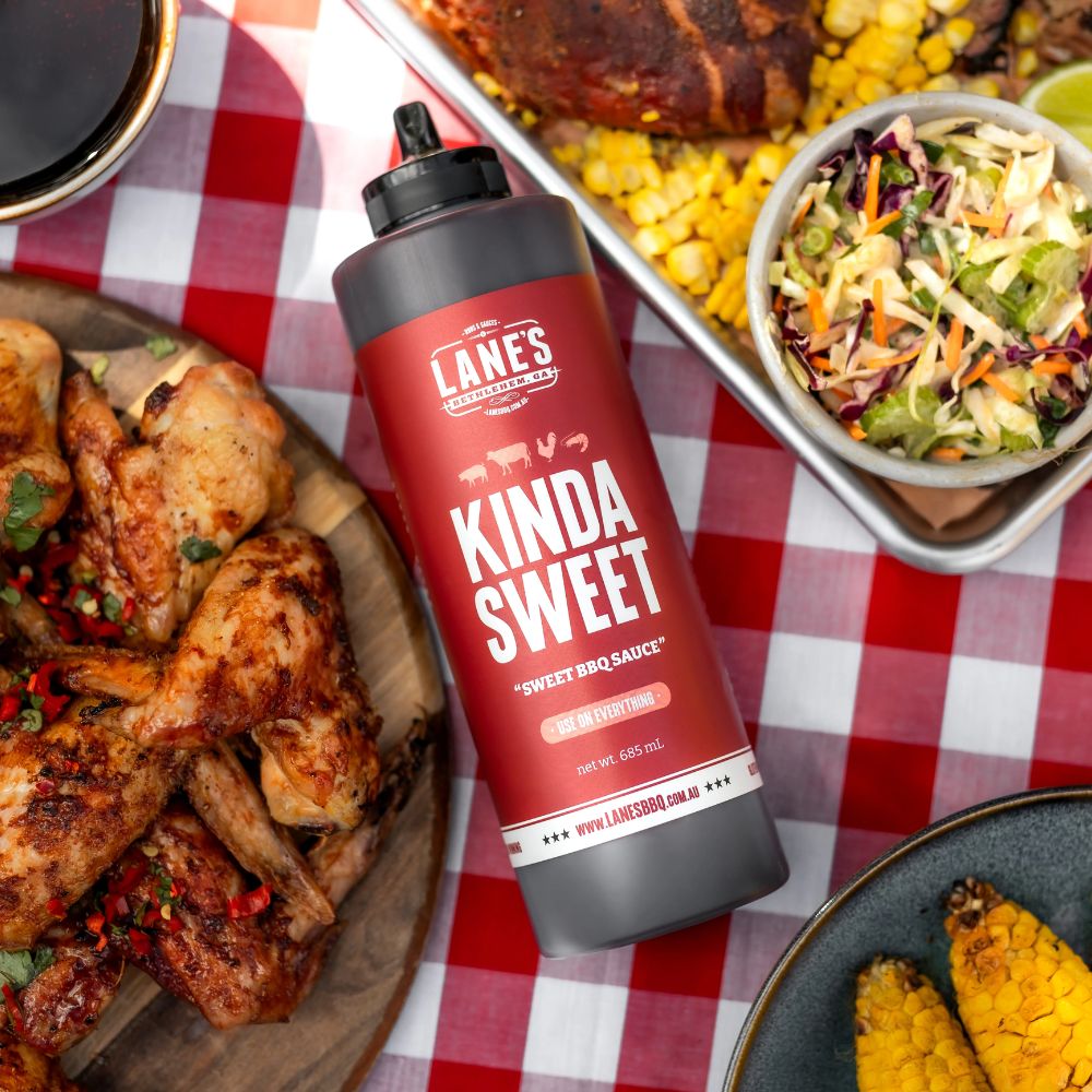 Lane’s BBQ Sauce “Kinda Sweet” Pitmaster bottle displayed on a red checkered table with grilled chicken wings, corn, and coleslaw, showcasing a sweet and tangy barbecue sauce served with classic BBQ sides.