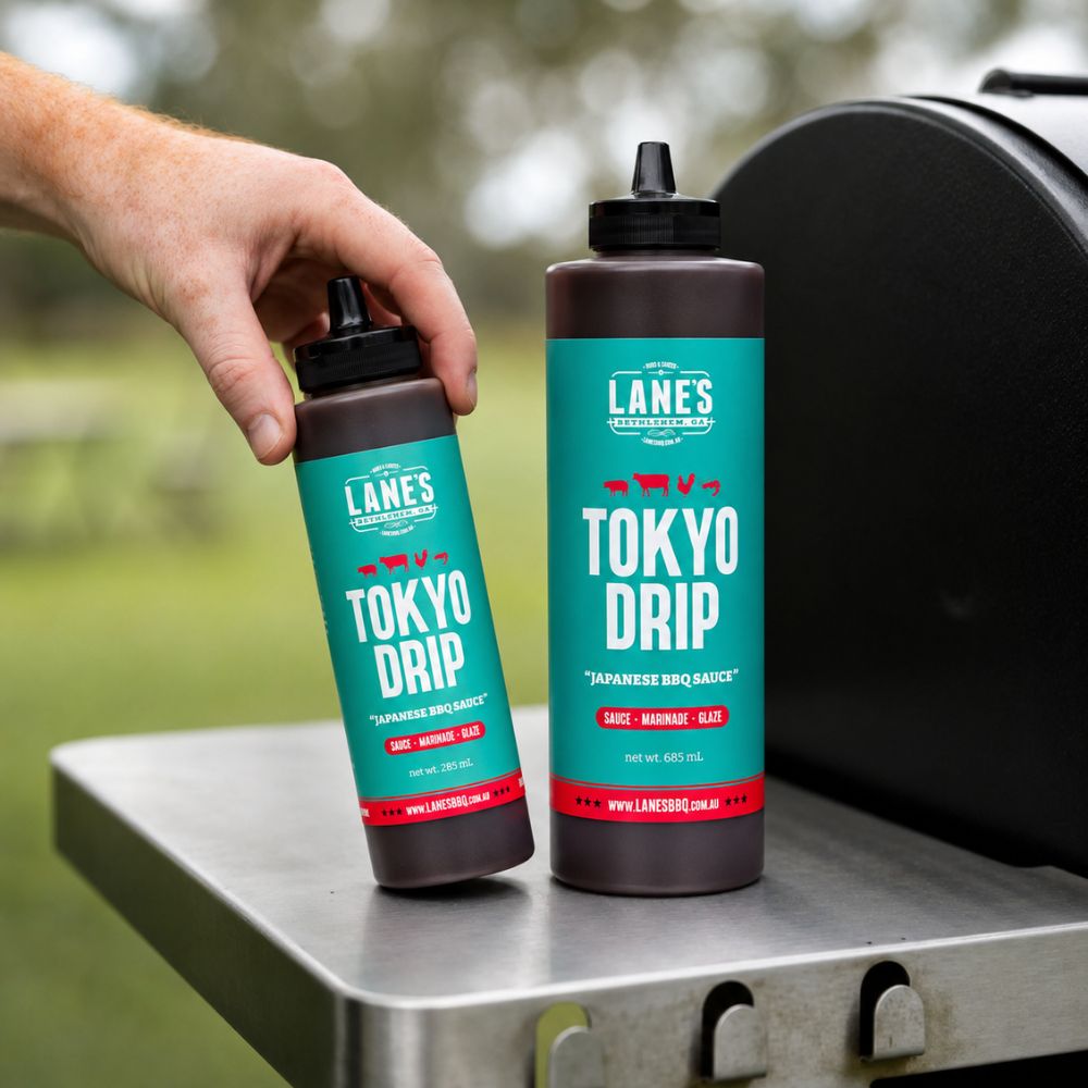 Lane's Tokyo Drip Sauce bottles in Small and Pitmaster sizes displayed beside a barbecue smoker, showcasing a Japanese-style BBQ sauce ideal for glazing, marinating, and finishing grilled meats.