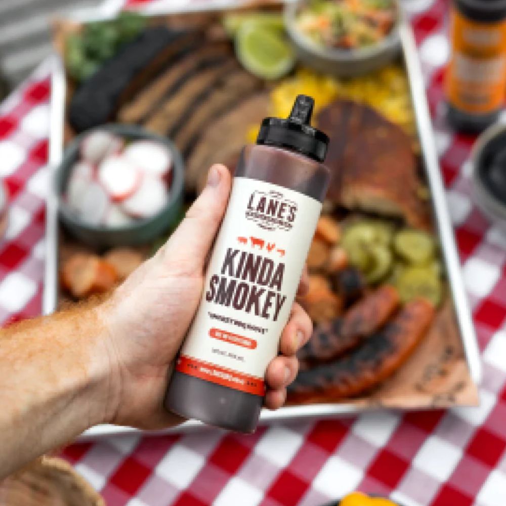Lanes BBQ Kinda Smokey pitmaster sauce bottle held over grilled meats and BBQ platter