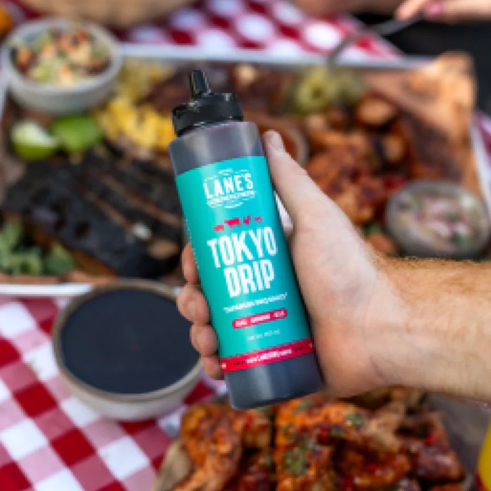 Lanes BBQ Tokyo Drip sauce pitmaster bottle held over grilled BBQ meats and sides