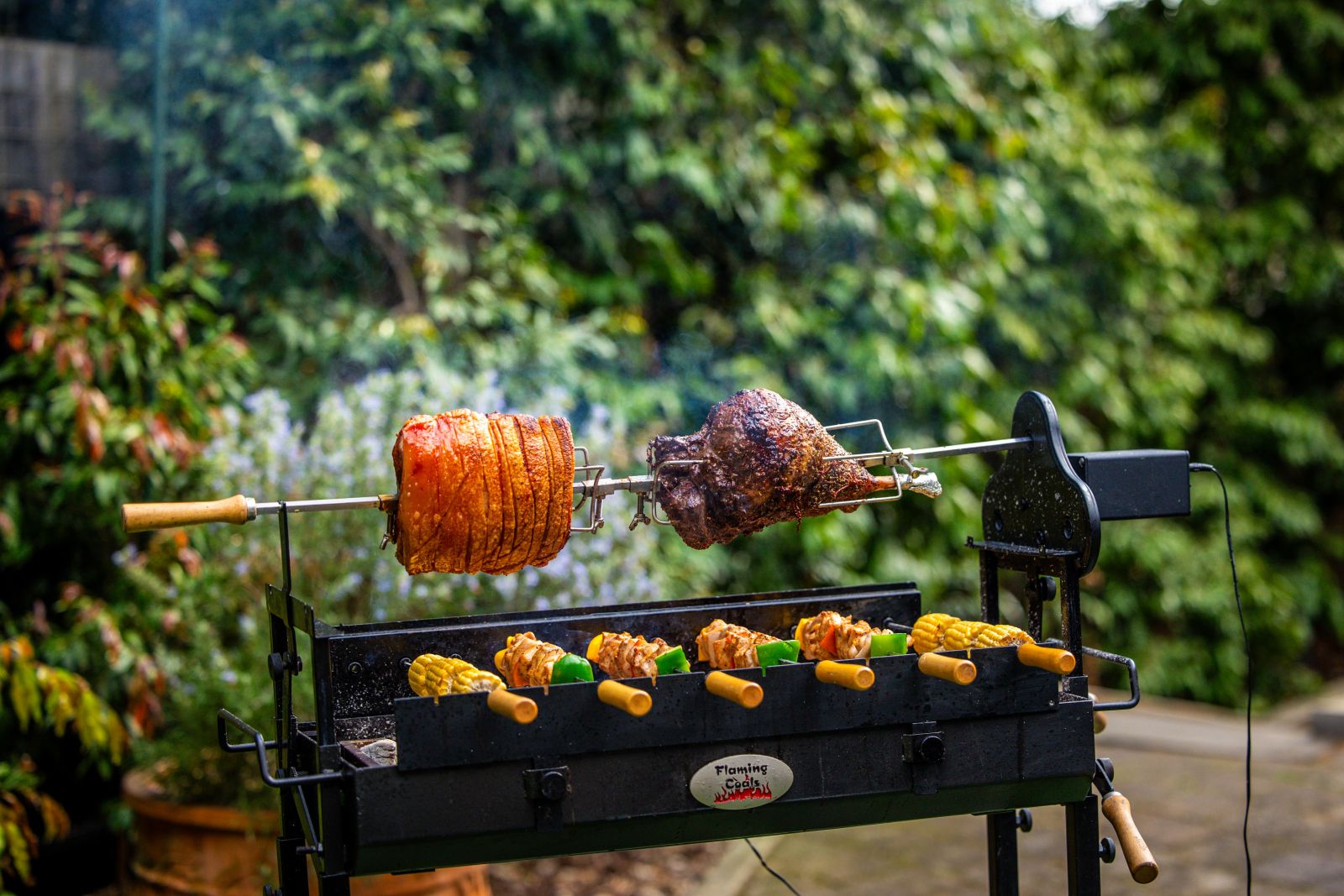Spit roasted pork shoulder and lamb cooking on a Flaming Coals Cyprus Spit with skewered vegetables over charcoal.