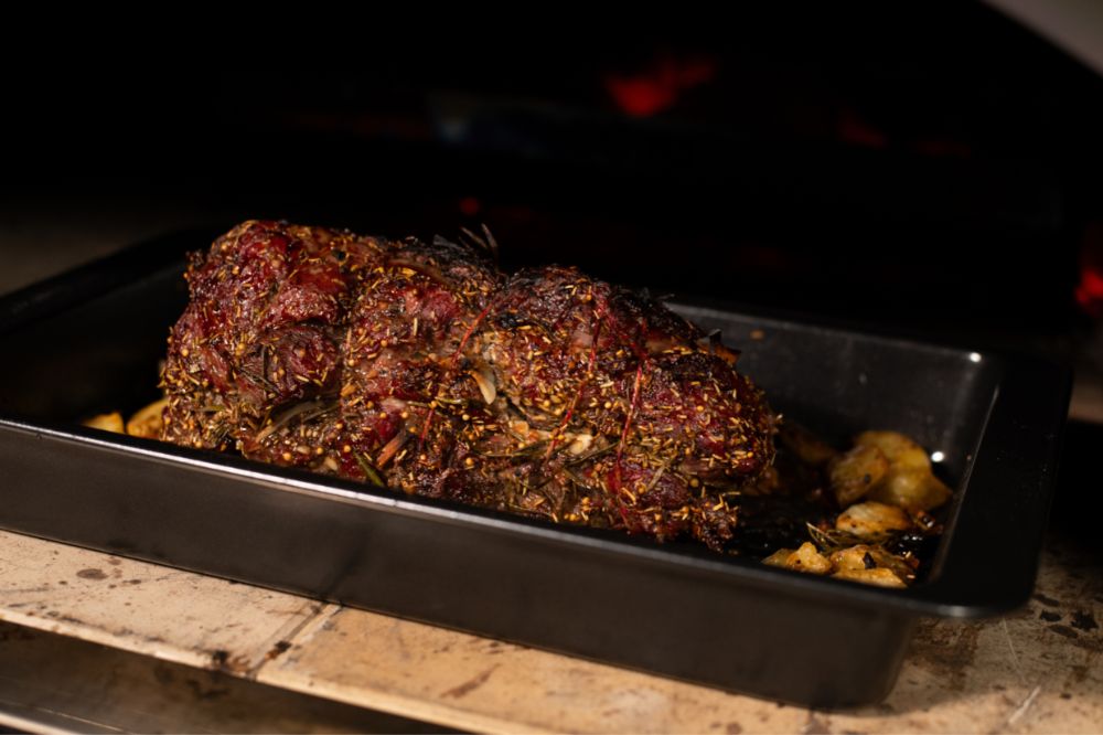 Herb-crusted lamb roast cooking in a black baking tray inside a wood-fired pizza oven, with golden roasted potatoes around the base.