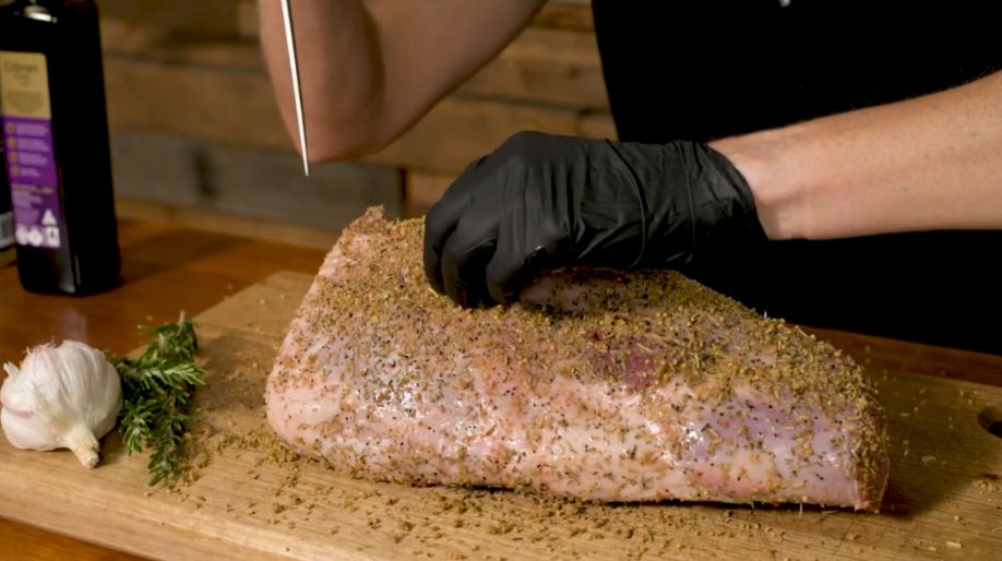 Hand wearing a black glove making small incisions in a seasoned leg of lamb, inserting garlic slithers and rosemary sprigs for extra flavour, on a wooden board.
